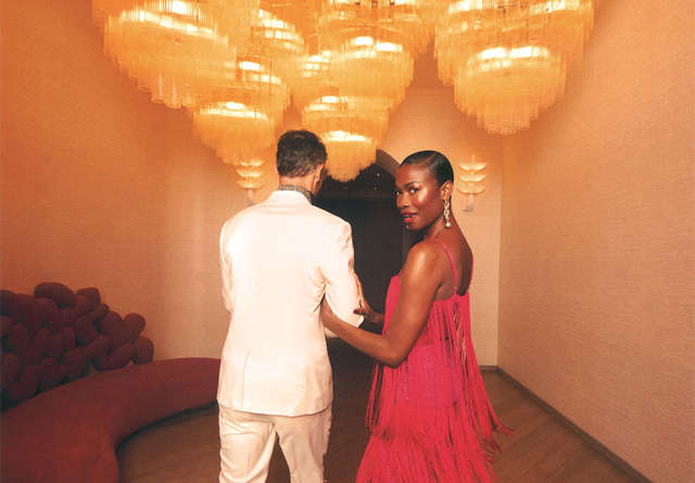 A couple walking through a stylish corridor in one of the Virgin Hotels in party attire and the woman glancing back